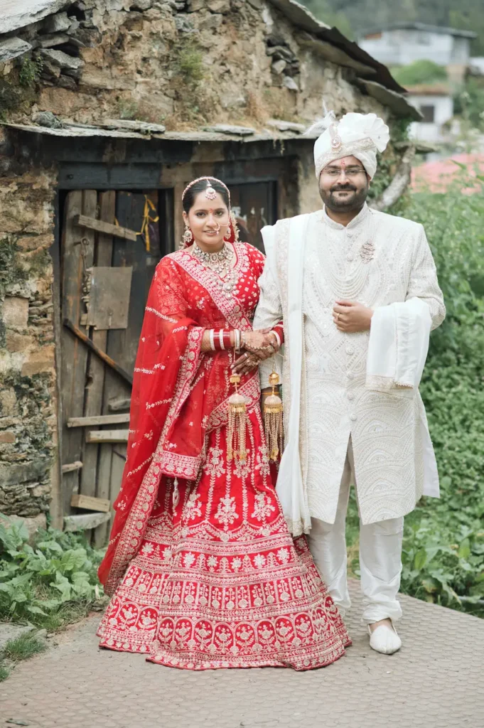 Bride and groom in traditional wedding attire posing together beside a rustic mountain house.