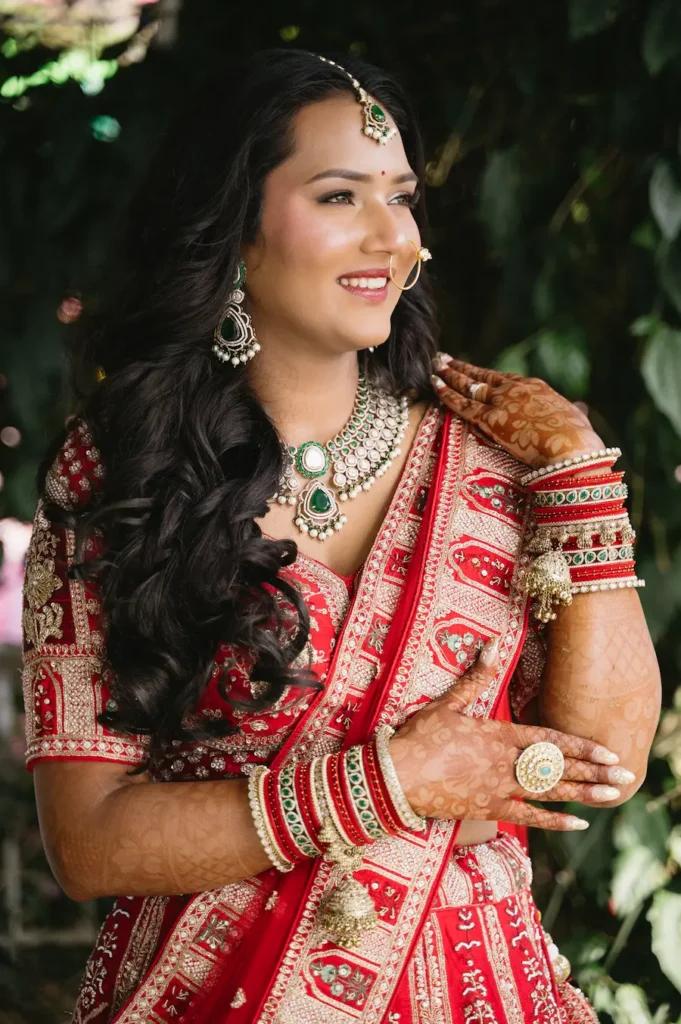 Smiling Indian bride in a red lehenga showing her beautiful mehndi and traditional emerald jewelry near Triyuginarayan Temple.