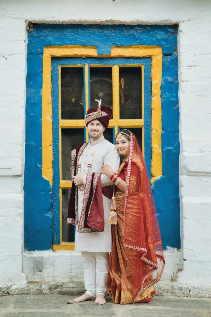 Bride and groom posing together in traditional wedding attire beside a blue and yellow window