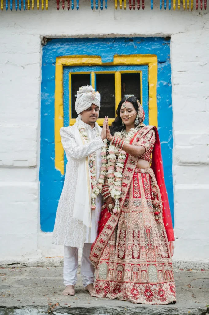 Smiling Indian bride and groom in traditional red and white wedding attire posing by a colorful blue and yellow window near Triyuginarayan Temple.