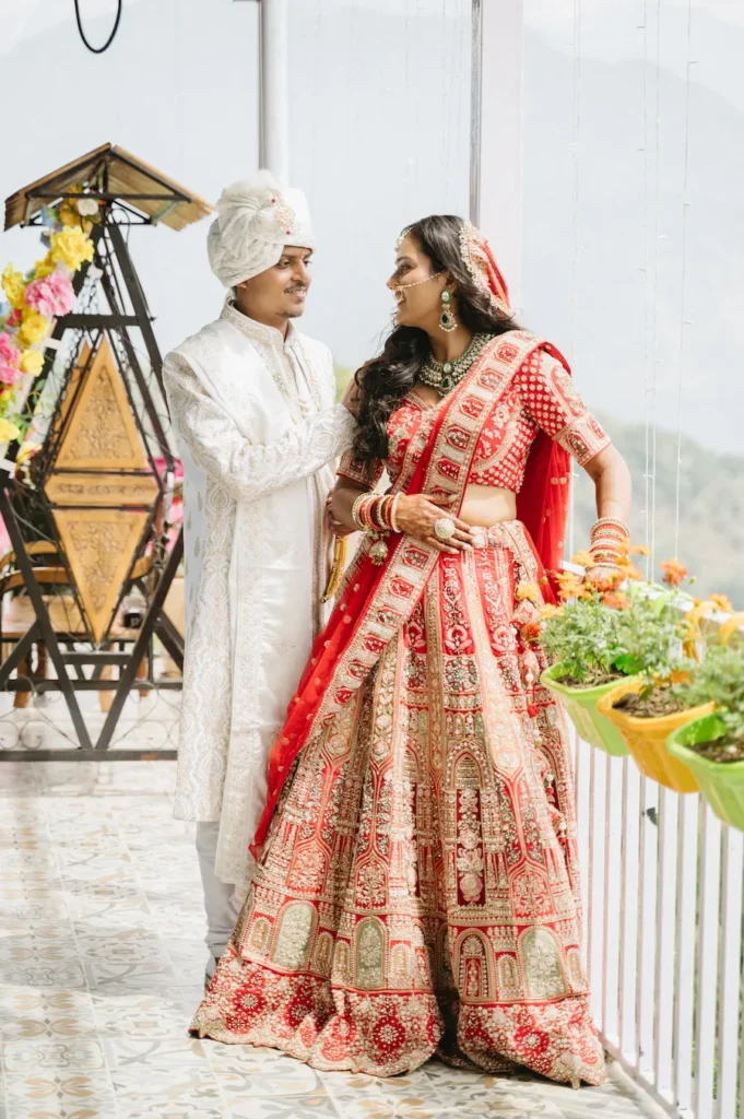 Indian bride in a red lehenga and groom in a white sherwani smiling at each other on a balcony near Triyuginarayan Temple.