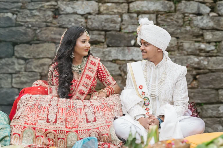 Bride and groom in traditional Indian attire sitting and smiling at each other by a stone wall near Triyuginarayan Temple.