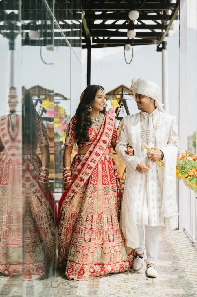 Smiling Indian bride and groom walking arm-in-arm along a bright corridor with a mirrored wall during their wedding near Triyuginarayan Temple.
