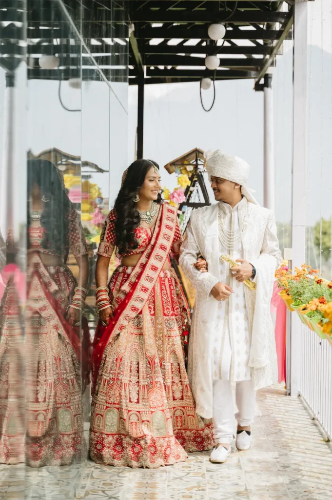 Smiling Indian bride and groom in traditional attire walking arm-in-arm along a bright corridor with a mirrored wall during their destination wedding in Dehradun.