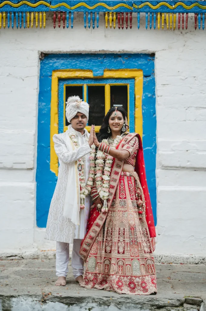 Smiling Indian bride and groom in traditional red and white attire playfully touching hands by a vibrant blue and yellow window near Triyuginarayan Temple.
