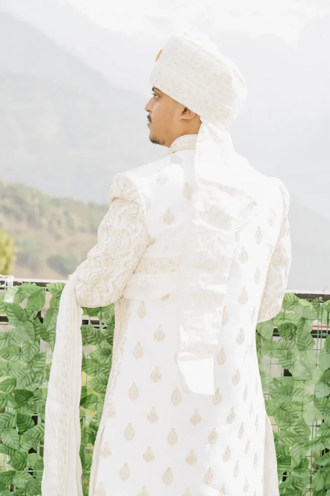 Indian groom in a white sherwani and turban looking out toward hazy mountains before his wedding ceremony.