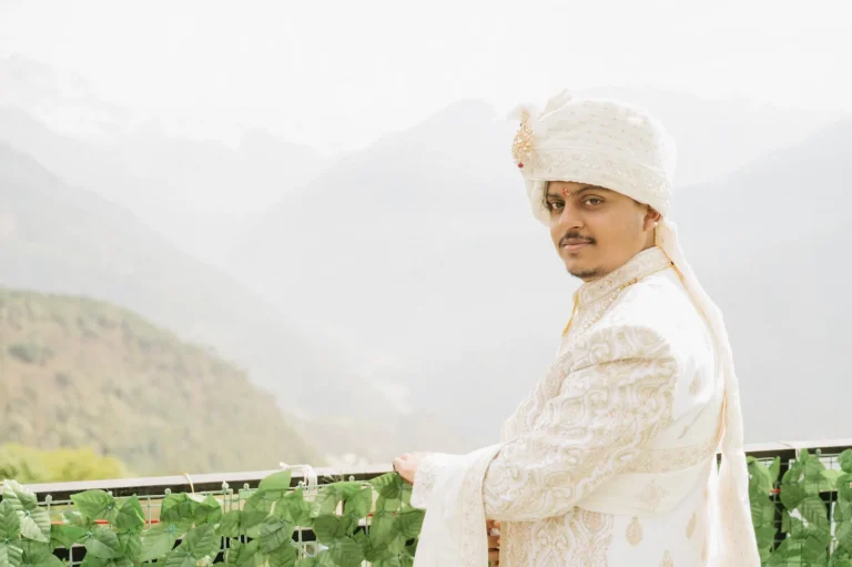 Indian groom in a white embroidered sherwani looking over his shoulder against a hazy mountain backdrop.