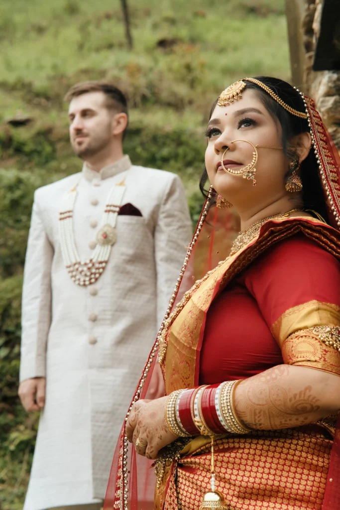 Bride in a red saree with the groom standing behind her in a lush mountain setting