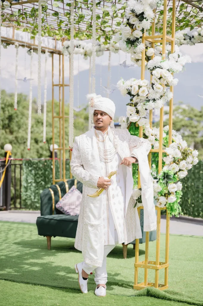 Indian groom in a white sherwani posing next to a gold and white floral wedding canopy.