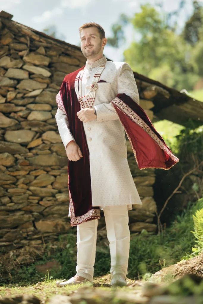 Groom wearing a traditional cream sherwani and maroon velvet shawl by a stone wall near Triyuginarayan Temple.