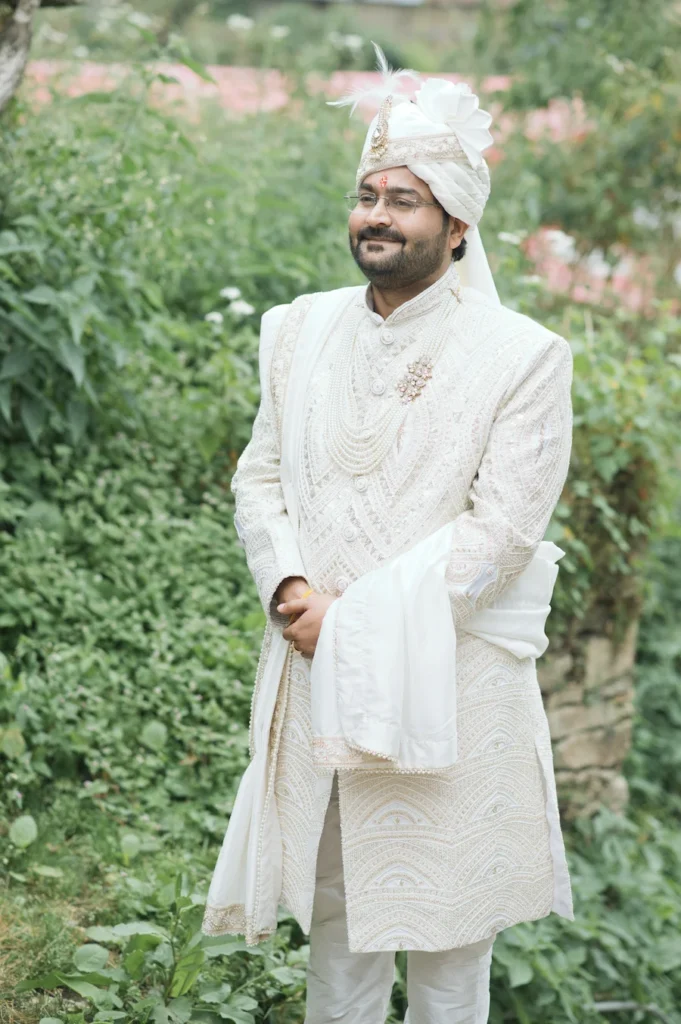 Groom in an elegant white sherwani and traditional turban posing outdoors surrounded by greenery.