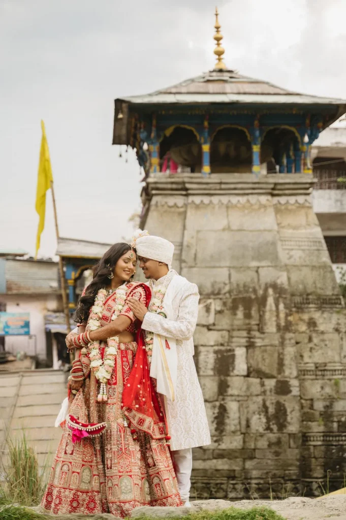 Indian bride and groom in traditional wedding attire embracing in front of an ancient stone temple in Triyuginarayan.