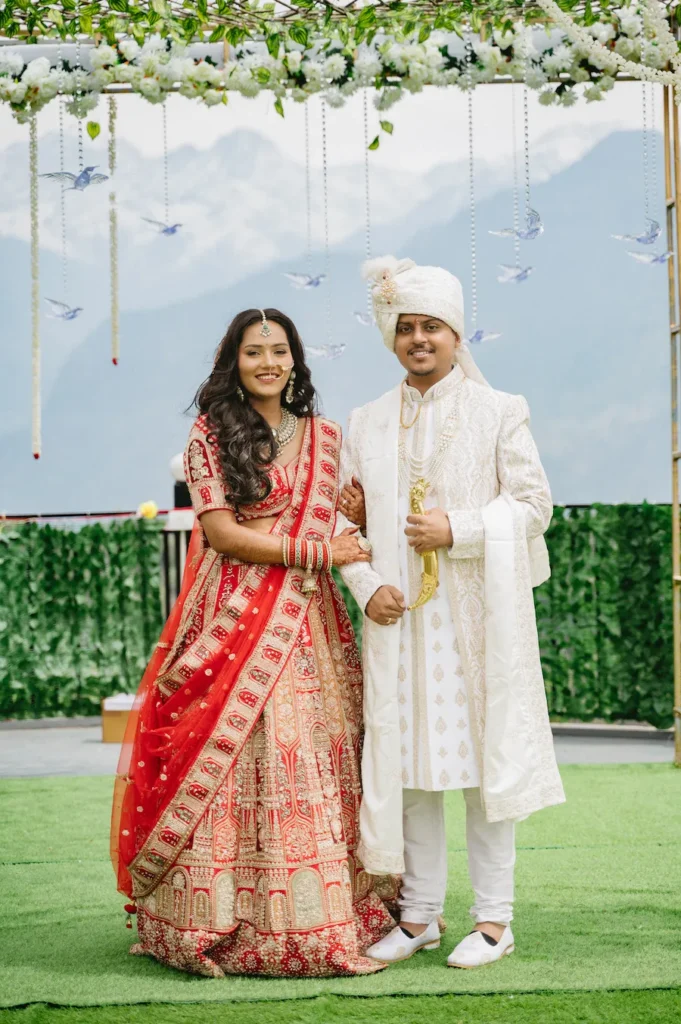 Smiling Indian bride in a red lehenga and groom in a white sherwani standing arm-in-arm under a floral canopy with a mountain backdrop.
