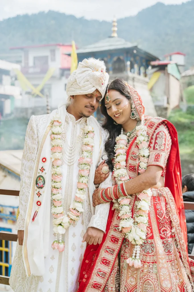 Indian newlywed couple wearing floral garlands sharing a quiet, affectionate moment with a temple and mountains in the background.