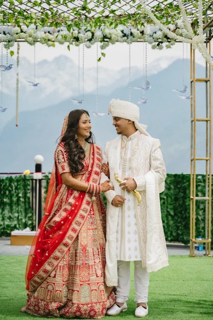 Smiling Indian bride and groom standing arm-in-arm looking at each other under a floral canopy with a mountain backdrop.