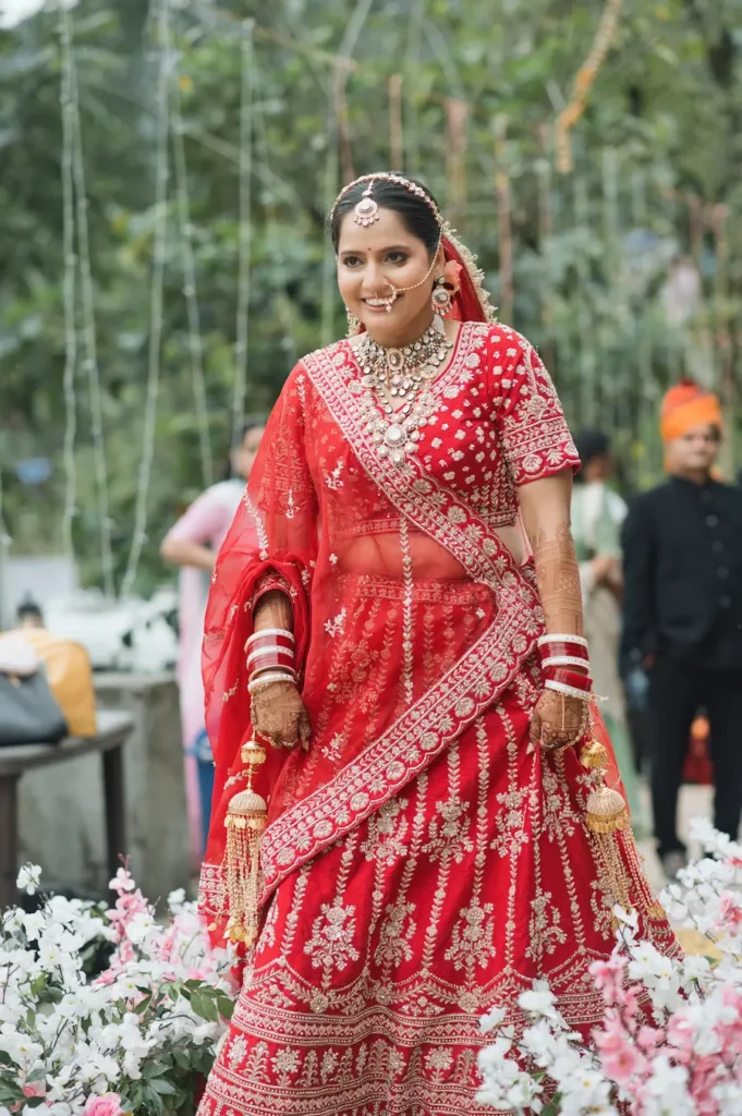 Bride in a traditional red bridal lehenga walking through a floral setting during a wedding.