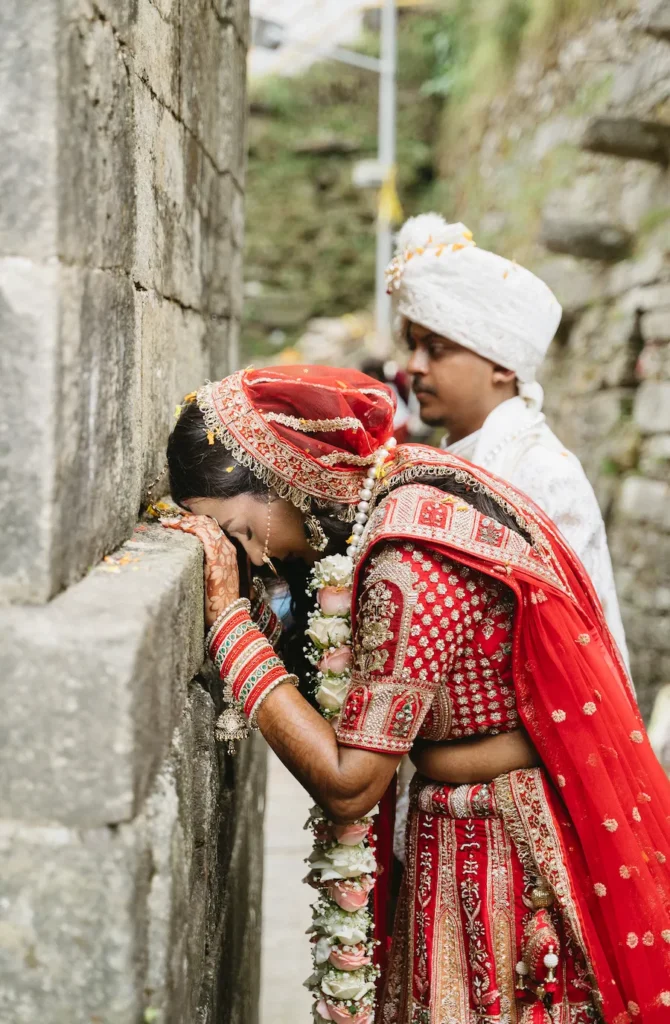 Indian bride in a red lehenga resting her head reverently against an ancient stone wall while her groom stands closely behind her.