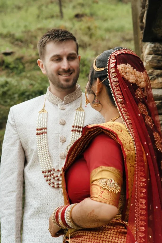 Smiling groom in a cream sherwani looking lovingly at his bride in a red saree near Triyuginarayan Temple.