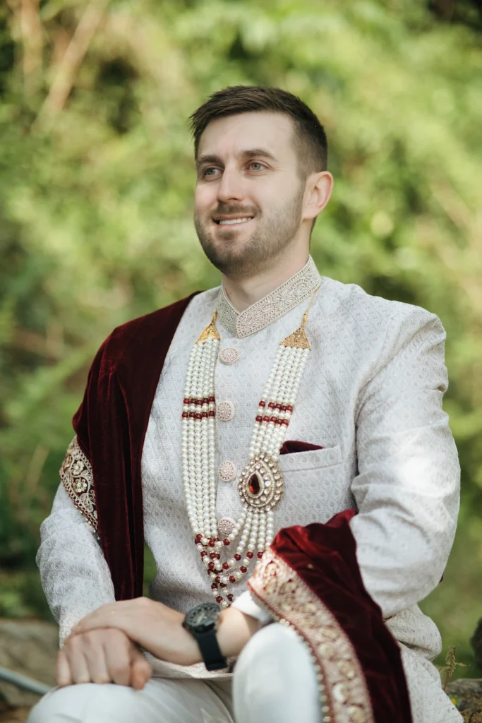 Smiling groom sitting outdoors in a traditional cream sherwani and maroon velvet shawl near Triyuginarayan Temple.