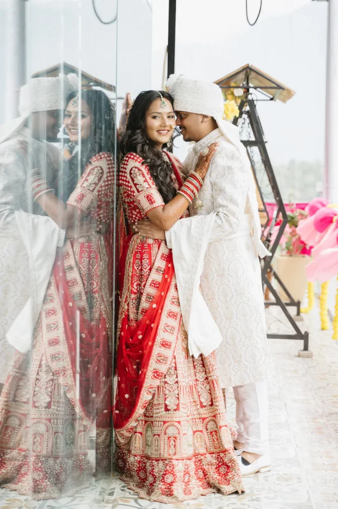 Smiling Indian bride and groom embracing next to a mirrored wall reflecting the bride's red lehenga during their Triyuginarayan temple wedding.