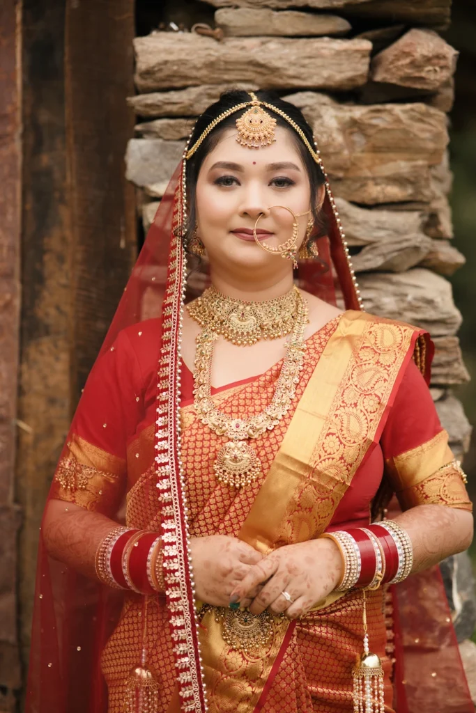 Traditional Indian bride wearing a red and gold saree and heavy gold jewelry by a stone wall near Triyuginarayan Temple.