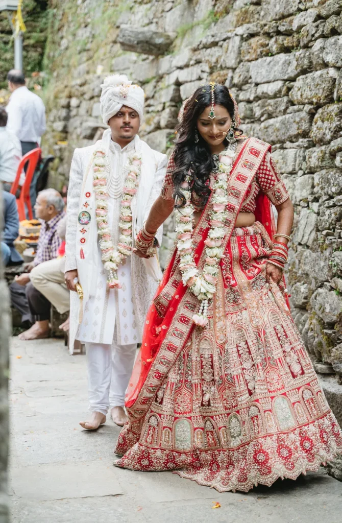 Indian bride in a red lehenga and groom in a white sherwani walking together along a stone wall during their traditional wedding ceremony.