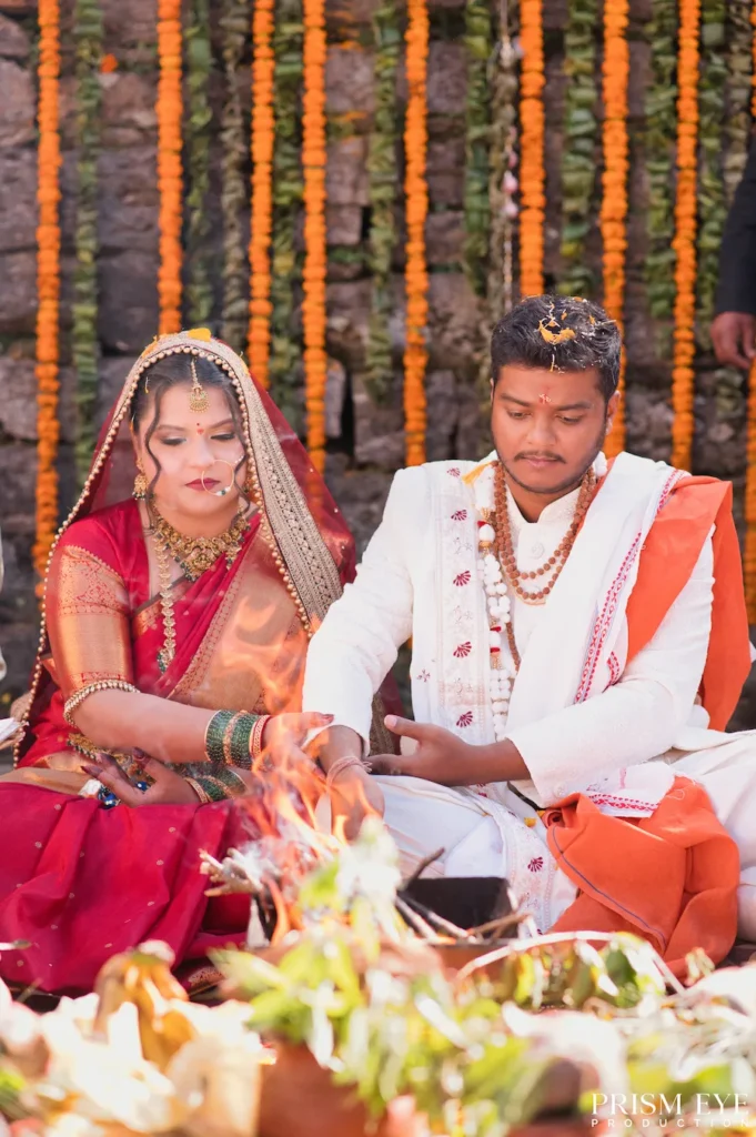 Indian bride in a red saree and groom in white attire performing a sacred fire ritual in front of a stone wall decorated with marigolds.