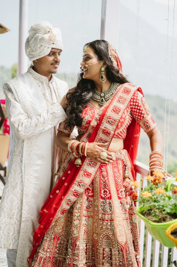 Smiling Indian bride in a red lehenga and groom in a white sherwani sharing a romantic glance on a scenic balcony near Triyuginarayan Temple.