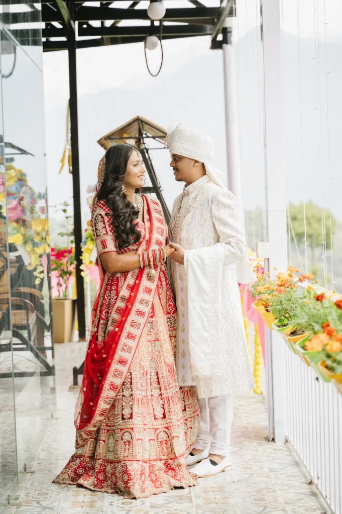Smiling Indian bride in a red lehenga and groom in a white sherwani holding hands and looking at each other on a balcony in Uttarakhand.