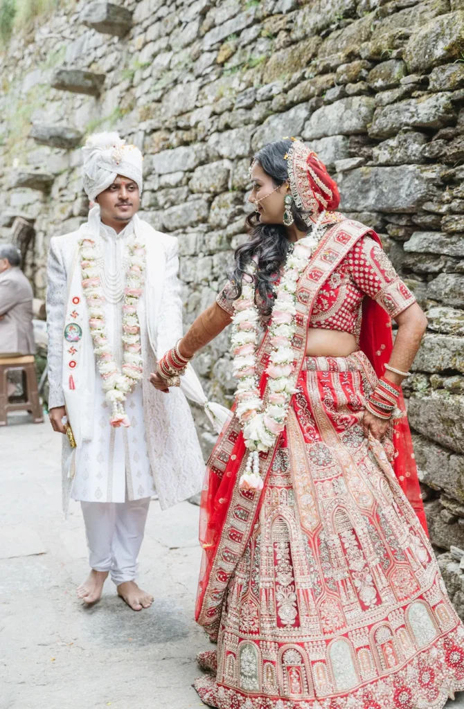 Smiling Indian bride in a red lehenga looking back at her groom in a white sherwani as they hold hands and walk barefoot along a stone path.