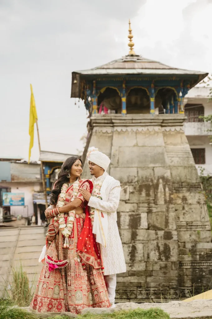Indian newlywed couple in traditional attire looking at each other affectionately in front of the stone Triyuginarayan Temple in Uttarakhand.