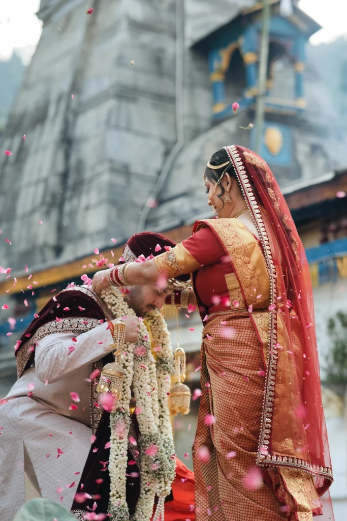 Bride placing a floral garland on the bowing groom under a shower of pink rose petals at Triyuginarayan Temple.