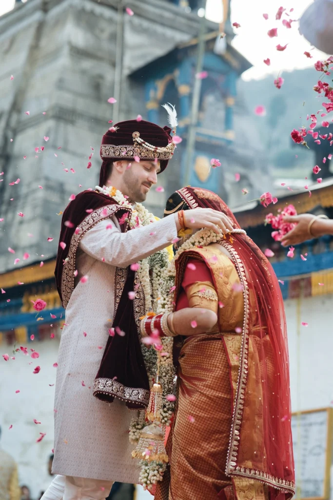 Groom placing a floral garland on the bride during their Varmala ceremony under a shower of pink petals at Triyuginarayan Temple.