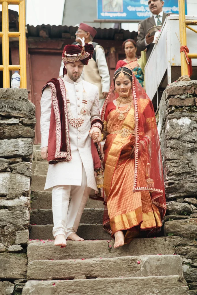 Bride and groom in traditional Indian wedding attire walking barefoot hand in hand down stone steps near Triyuginarayan Temple.