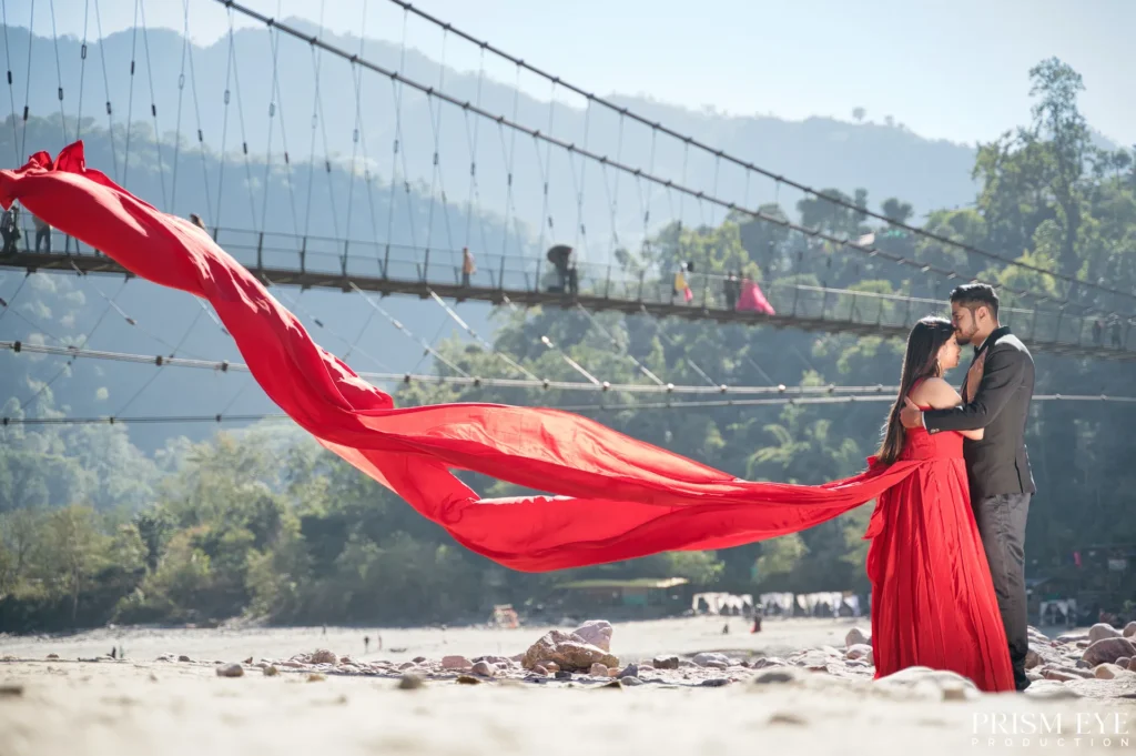 Couple embracing on a Ganga beach in Rishikesh, with the bride's flowing red dress creating a dramatic romantic moment beneath the suspension bridge.