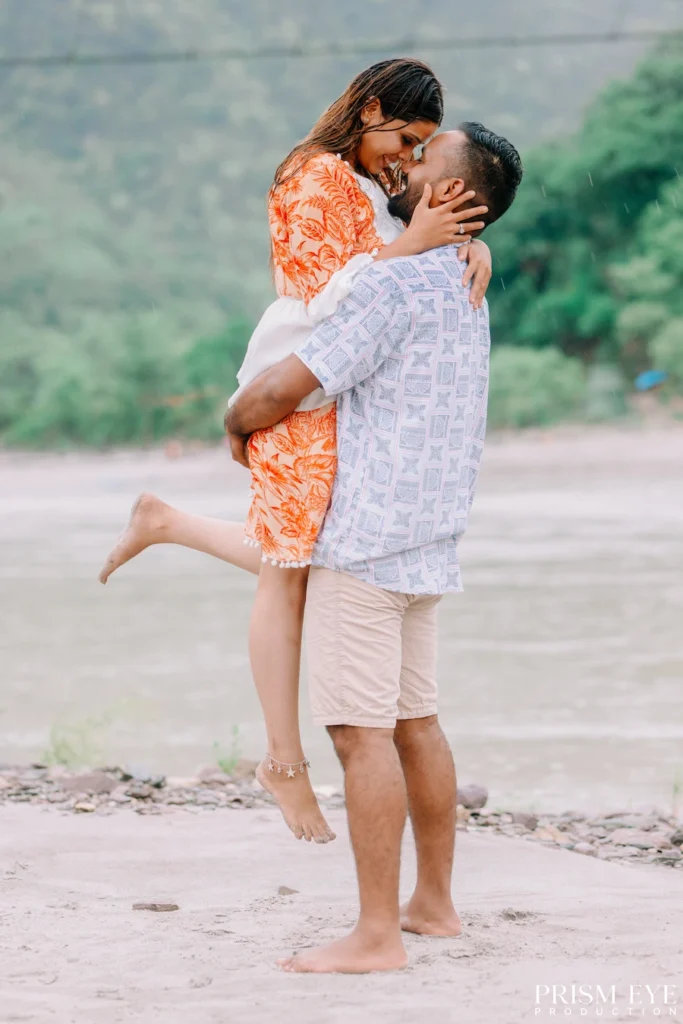 A couple sharing a joyful embrace on the Ganga beach in Rishikesh, with the bride lifting her leg as they celebrate their love.