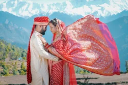 A breathtaking destination wedding portrait where the bride's flowing dupatta adds movement and elegance against the stunning Himalayan backdrop. Bride and groom embracing with a flowing bridal dupatta against the Himalayan mountains during their destination wedding in Uttarakhand.
