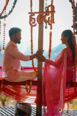 A joyful Haldi ceremony moment as the couple smiles at each other while seated on a beautifully decorated swing surrounded by marigold flowers. Couple sharing a candid moment on a decorated swing during their Haldi ceremony with marigold floral decorations.