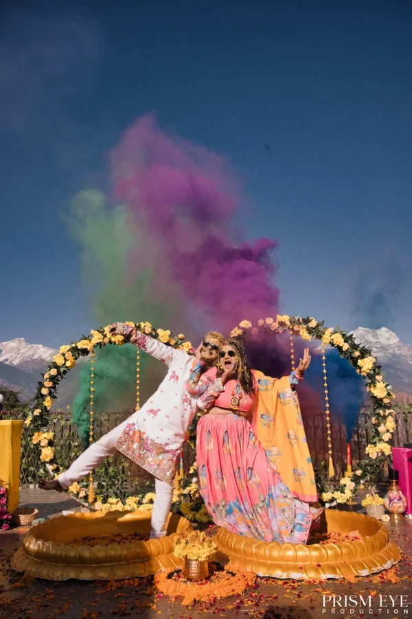 Couple celebrating with colorful Holi powder beneath a floral arch with Himalayan peaks in the background