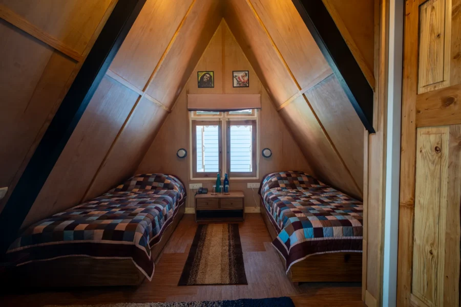 Warm and cozy A-frame wooden bedroom in Dehradun featuring twin beds, natural wood interiors, and a peaceful mountain retreat atmosphere. Cozy wooden A-frame bedroom with twin beds in a luxury cabin in Dehradun, Uttarakhand.