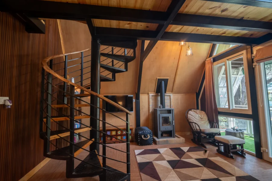 Elegant A-frame cabin interior in Dehradun featuring a spiral staircase, wooden finishes, and a cozy luxury mountain retreat ambiance. Luxury wooden A-frame cabin interior with spiral staircase and large windows in Dehradun, Uttarakhand.