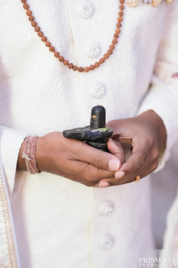 Close-up of an Indian groom's hands holding a black Shiva Lingam idol against his white sherwani during a wedding ritual.