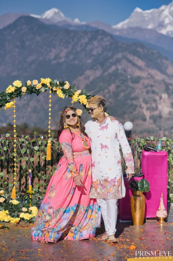 Smiling couple wearing sunglasses and covered in yellow haldi paste posing in front of a floral arch with the Chaukhamba mountain range in the background.