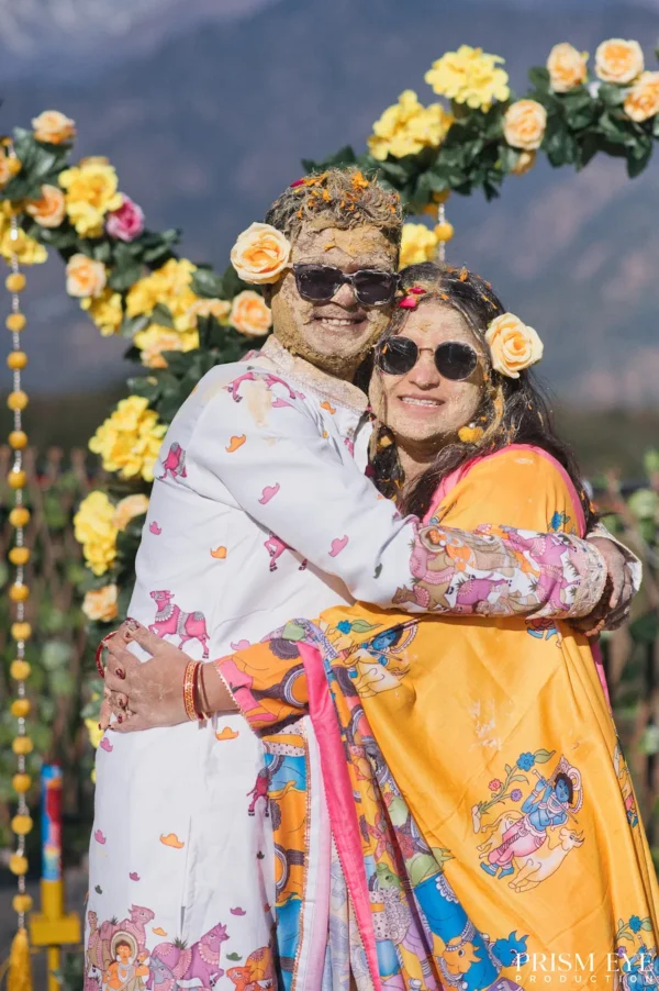 Joyful couple wearing sunglasses and covered in yellow haldi paste hugging outdoors with a floral arch and mountains in the background.