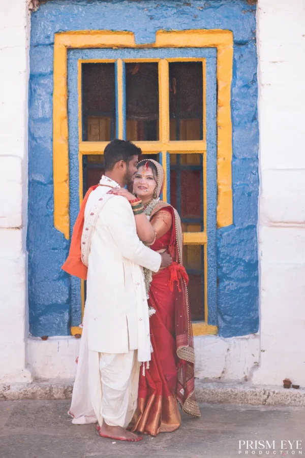 Smiling Indian bride in a red saree and groom in a white dhoti kurta embracing in front of a brightly painted blue and yellow window.
