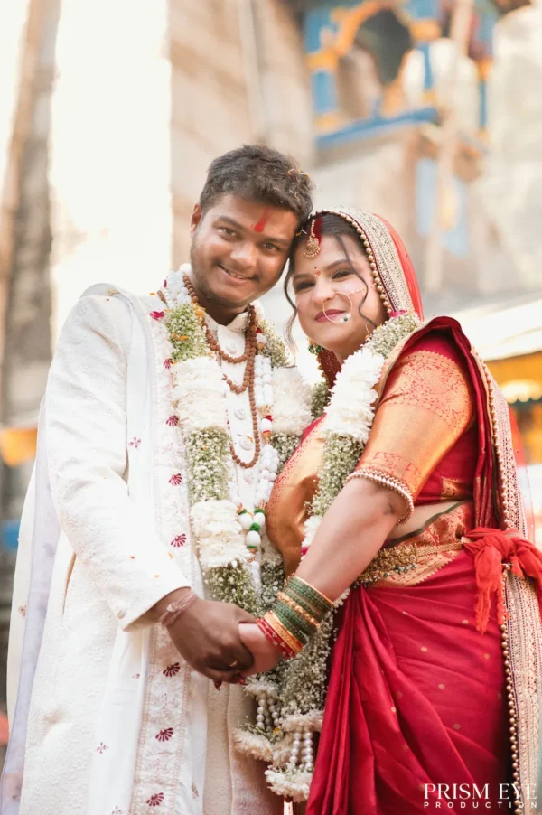 Smiling Indian newlywed couple wearing floral garlands and traditional attire holding hands and looking at the camera.