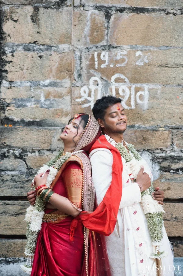 Indian bride and groom standing back-to-back with eyes closed, leaning against a rustic stone wall with Hindi inscriptions.